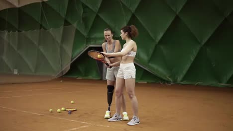 A-beautiful-female-coach-with-a-prosthesis-on-her-leg-shows-her-ward-the-nuances-of-the-ball-hitting-in-tennis-standing-behind-her.-Sports-girls