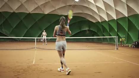 Rückseitenaufnahmen-Von-Zwei-Athletischen-Mädchen,-Die-Auf-Einem-überdachten-Tennisplatz-Tennis-Spielen.-Frau-Im-Rahmen-Mit-Einer-Beinprothese