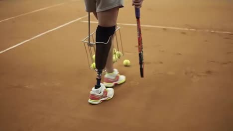 Tracking-footage-of-a-sports-girl-with-a-prosthesis-on-her-right-foot-picking-up-tennis-balls-with-the-basket.-Tennis-player.-Back-view