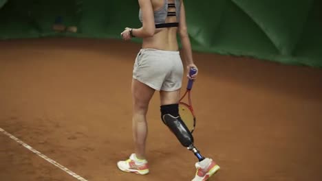 Sporty-disable-girl-taking-a-ball-from-a-basket,-walk-to-the-middle-and-make-a-strong-shot-with-the-tennis-racket.-Indoors
