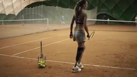 Two-beautiful-girls-practicing.-The-handicapped-girl-in-short-shorts-takes-the-ball-from-the-basket-and-makes-the-first-serve-of-the-ball.-Indoor-tennis-court.-Back-view