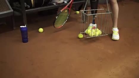 Athlete,-disabled-female-collects-tennis-balls-in-the-basket-helping-herself-with-a-racket.-Close-up