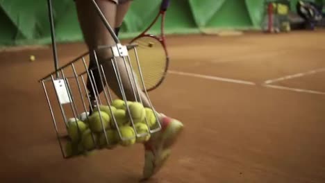 Aiming-footage-of-a-disabled-woman's-legs.-Slender-girl-collects-balls-with-a-tennis-basket.-Indoors-tennis-court