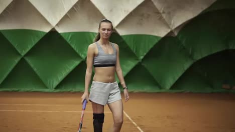 Young-long-haired-woman-in-sportswear-is-walking-on-the-indoor-tennis-court-with-racket-in-her-hand.-Green-tent-behind.-Slow-motion