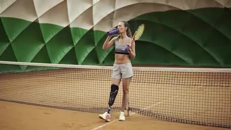 Seductive-tennis-girl-with-the-leg-prosthesis-is-standing-near-the-net-on-the-tennis-court,-drinking-water,-smacks-sweat-from-the-forehead.-In-full-length