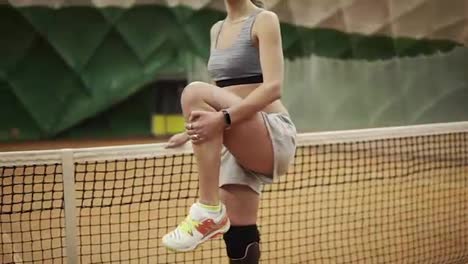 A-handicapped-woman-leaning-on-a-tennis-net-is-standing-on-one-leg,-warm-up-her-ankle.-High-spirited.-Indoors
