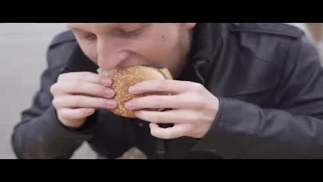 Young-man-with-beard-in-the-street-cafe-biting-tasty-big-burger-with-cheese.-French-fries-on-the-plate.-Shot-in-4k
