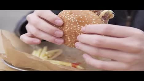 Closeup-view-of-man's-hands-holding-big-tasty-burger.-Man-with-beard-in-the-street-cafe-biting-tasty-big-burger-with-cheese.-French-fries-on-the-plate.-Shot-in-4k