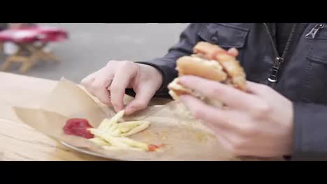 Young-man-with-beard-in-the-street-cafe-eating-french-fries-and-biting-tasty-big-burger-with-cheese.-Shot-in-4k