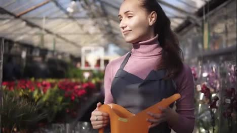 Young-female-gardener-in-uniform-walking-in-a-greenhouse-among-shelves-with-different-plants-and-holding-orange-watering-can-in-her-hands