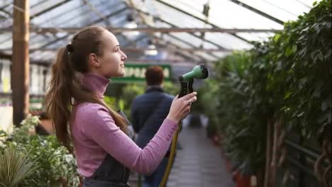 Young-attractive-female-gardener-in-uniform-watering-plants-with-garden-hose-in-greenhouse.-Slowmotion-shot