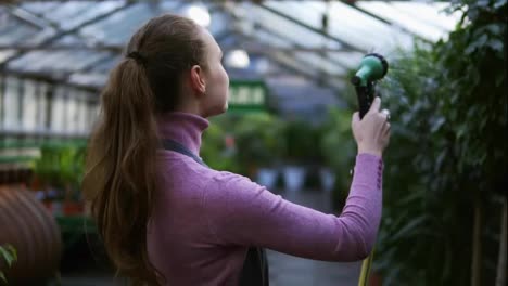Young-smiling-female-gardener-in-uniform-watering-plants-with-garden-hose-in-greenhouse.-Slowmotion-shot