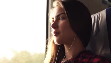 Side-view-of-a-beautiful-caucasian-girl-going-somewhere-by-train.-Listen-music-in-earphones.-Pensive-female.-Smiling.-Sun-shines