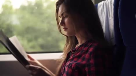 Young,-pretty-girl-travelling-by-modern-train.-Sitting-next-to-the-window-and-reading-in-plaid-shirt.-Side-view.-Blurred-window-in-motion