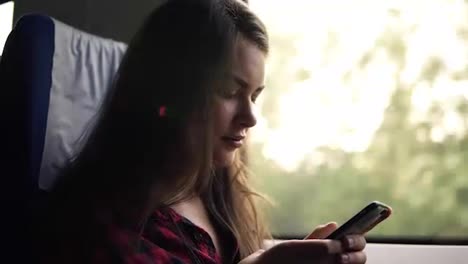 Low-angle-side-view-of-a-beautiful-girl-typing-in-her-mobile-phone.-Going-by-train.-Travelling.-Nature-landscape-in-window.-Motion