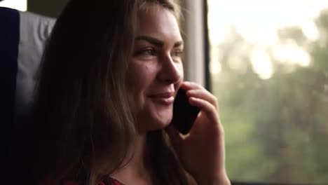 Brown-haired-young-girl-travels-by-modern-train.-Next-to-the-window.-Speaks-on-the-mobile-phone.-Smiling.-Daytime
