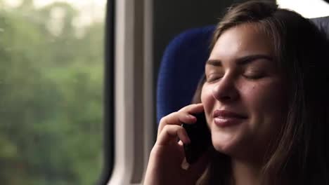 Close-up-of-woman's-face.-Pretty-girl-is-travelling-by-train.-Talking-on-her-mobile-phone.-Smiling,-relaxed.-Moving-picture-of-nature-in-the-window