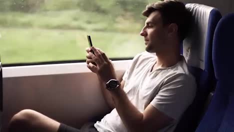 Side-view-of-a-young,-handsome-man-travels-by-modern-train.-Sitting-next-to-the-window-and-looking-at-his-mobile-phone.-Slight-natural-train-shaking.-Casual-clothes.-Nature-landscape-in-window