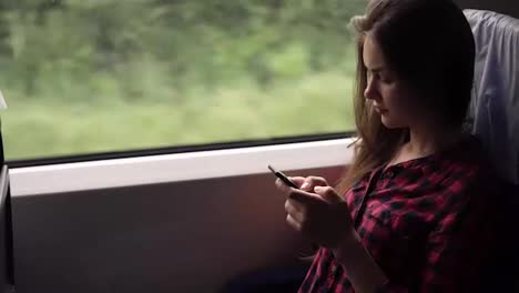 Serious,-relaxed-young-girl-sitting-in-the-train-near-the-window.-Travelling-by-modern-train.-Long-haired-girl-is-scrolling-or-typing-on-her-mobile-phone.-Plaid-red-shirt.-Side-view
