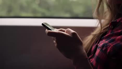 Extremely-close-up-of-woman's-hands-with-mobile-phone.-Casual-plaid-shirt.-Pretty-girl-is-travelling-by-train.-Nature-landscape-in-window-in-motion.-Side-view