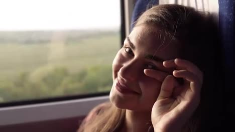 Attractive,-young-girl-is-smiling-at-the-camera.-Traveling-by-train.-Green-nature-outside-the-window.-The-girl-looks-thoughtfully-out-the-window.-Close-up