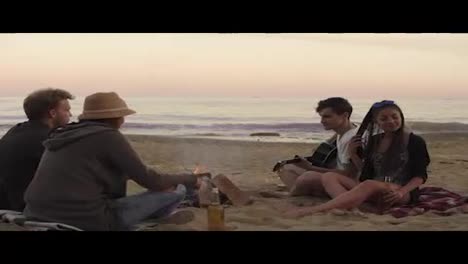 Young-friends-sitting-by-the-fire-on-the-beach-in-the-evening,-drinking-beer-and-playing-guitar.-Shot-in-4k