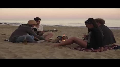 Young-cheerful-friends-sitting-by-the-fire-on-the-beach-in-the-evening,-drinking-beer-and-playing-guitar.-Shot-in-4k