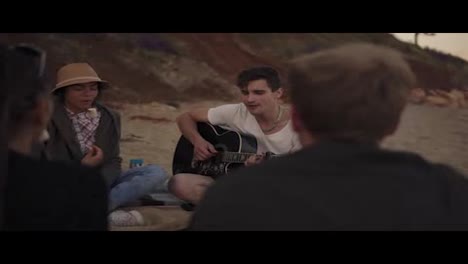 Young-cheerful-friends-sitting-by-the-fire-on-the-beach-in-the-evening,-cooking-marshmallow-and-playing-guitar.-Shot-in-4k