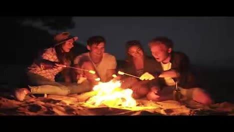Young-cheerful-friends-sitting-by-the-fire-on-the-beach-in-the-evening,-cooking-marshmallow-on-sticks-together.-Shot-in-4k