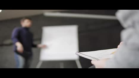 Closeup-view-of-woman-taking-notes-during-the-presentation.-Handsome-young-businessman-pointing-at-flipchart-during-presentation-in-conference-room.-Shot-in-4k