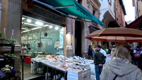 People-and-sellers-in-Via-Pescherie-Vecchie,-a-famous-alley-full-of-traditional-stores-and-food-stalls-in-the-characteristic-medieval-city-centre-of-Bologna,-Italy