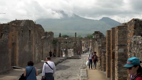 Tourists-Walking-Along-Pompeii-Ruins-On-Via-Della-Scuole-With-Mount-Vesuvius-Covered-In-Clouds-In-Background