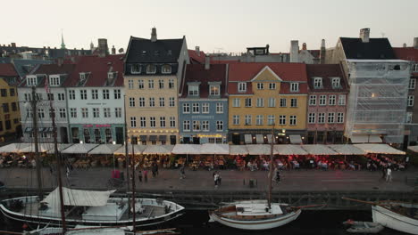 A-picturesque-sunset-over-Nyhavn,-Copenhagen,-setting-a-cozy-mood-with-lively-crowds-and-boats-sailing-along-the-canal