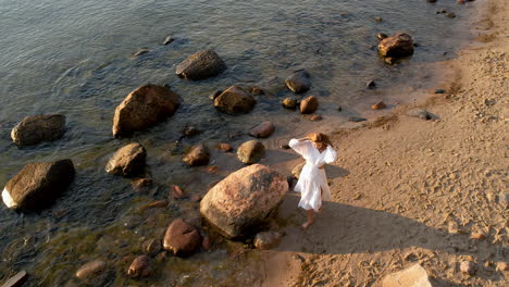 A-woman-walks-along-the-seashore-during-sunrise-dressed-in-a-flowing-white-dress---view-from-above-from-a-drone