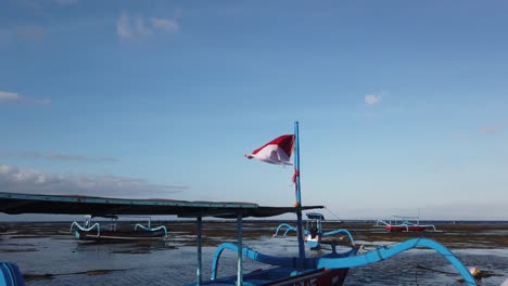 National-Flag-of-Indonesia-Waves-in-the-Wind-at-Boats-Seascape,-Sanur-Bali-Beach