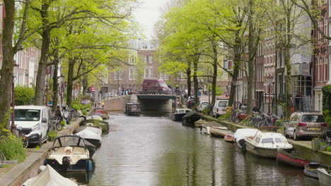 A-picturesque-view-of-an-Amsterdam-canal,-bordered-by-lush-green-trees,-with-a-boat-traversing-the-water