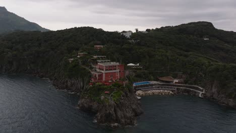 Hotel-Mezzatorre-and-forested-coastal-cliffs-at-Ischia,-aerial-push-in