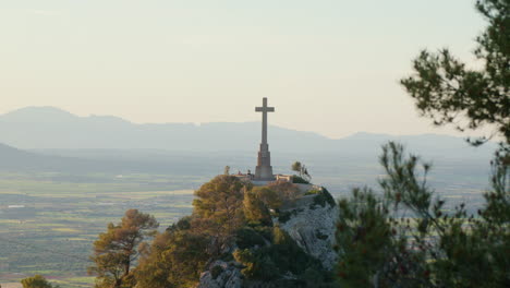 A-hill-at-the-Santuari-de-San-Salvador-in-Mallorca,-Spain,-crowned-by-a-cross,-with-a-mountain-range-forming-a-majestic-backdrop-in-the-late-afternoon