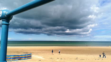 Ominous-storm-cloud-approaching-golden-sandy-Anglesey-beach,-looking-through-metal-railings