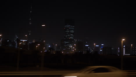 Driving-in-a-car-and-passing-by-Dubai-skyline-at-night-in-a-car-on-the-highway-with-distant,-lit-up-buildings-and-skyscrapers-including-the-Burj-Khalifa-in-the-United-Arab-Emirates