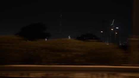 Passing-by-Dubai-skyline-at-night-in-a-car-on-the-highway-with-distant,-lit-up-buildings-and-skyscrapers-including-the-Burj-Khalifa-in-the-United-Arab-Emirates