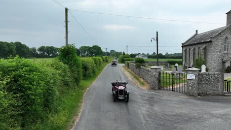 A-group-of-vintage-cars-on-a-country-lane-passing-a-church-on-a-classic-car-rally-event-in-Carlow-Ireland-on-a-bright-summer-morning