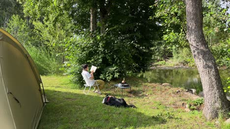 An-Asian-girl-camping-and-reading-alone-by-the-river,-with-her-dog