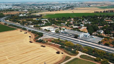 A-drone-shot-flying-above-a-huge-yellow-green-field,-with-cars-driving-on-a-road-towards-a-cityscape-full-of-buildings,-4K-video,-Israel