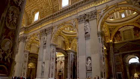 An-establishing-shot-of-the-ceiling-of-the-Saint-Peter's-Basilica-covered-in-mosaics,-tilting-down-to-reveal-the-vast-open-space-and-tourist-going-about-their-day-in-the-Vatican-City,-Italy
