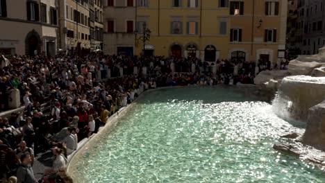 An-establishing-shot-of-a-crowd-of-tourist-gathered-around-the-Trevi-Fountain,-a-slow-pan-to-reveal-the-beautiful-historical-fountain-on-a-sunny-day,-Rome,-Italy