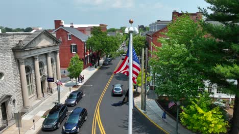 Drone-footage-of-a-downtown-area-of-a-small-town-with-municipal-buildings,-people-walking-down-the-sidewalks-and-an-American-flag-with-security-cameras-mounted-on-top,-gently-blowing-in-the-breeze