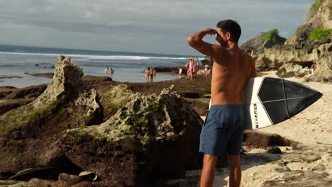 A-handsome-young-man-looking-a-waves-with-surfboard-in-his-hand-on-a-sunny-day-on-Suluban-beach-Bali-Indonesia