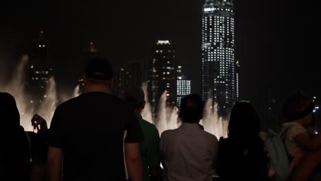 Crowd-of-people-watching-the-water-fountain,-light-show-at-the-Dubai-mall-with-lit-up-skyscrapers-in-distance