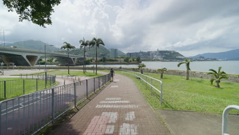 Walking-along-brick-pathway-above-river-with-highway-and-buildings-of-Hong-Kong-in-distance
